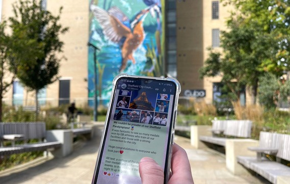 A hand holding a phone on a sunny day in Pounds Park, the screen shows Sheffield City Council's WhatsApp Channel. 