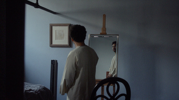 A Black man in a dimly lit room, looking at his reflection in a mirror