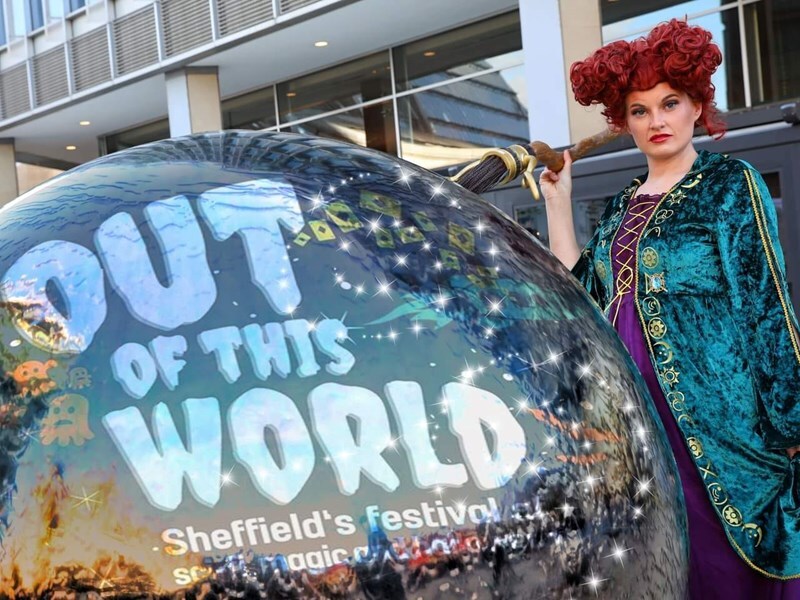 Out of this World imposed on a large shiny ball, with a women dressed in fancy dress with a red wig standing beside it.