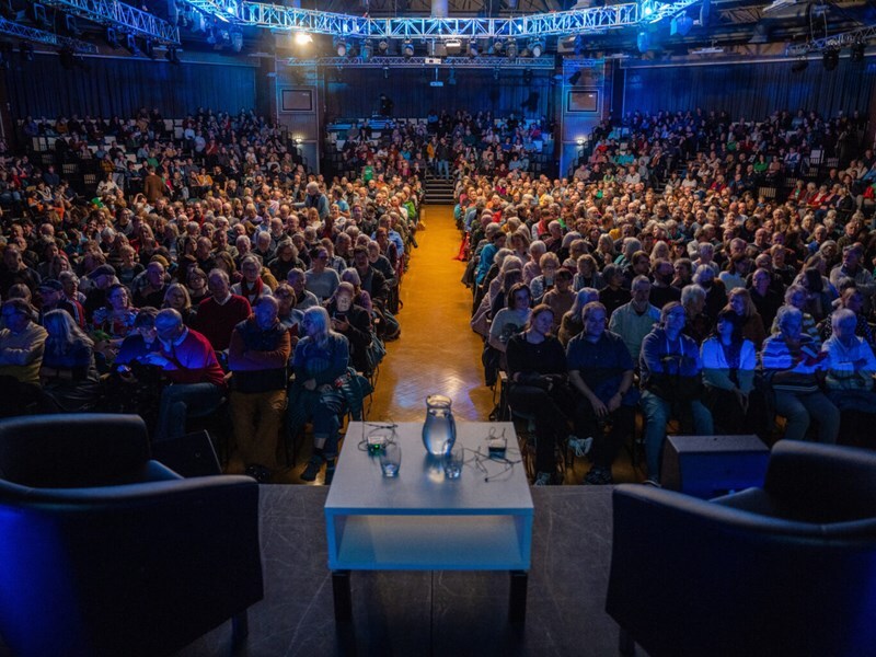 A packed out audience sitting down, looks towards a stage with a table in between two chairs, ready for the speakers to come out.