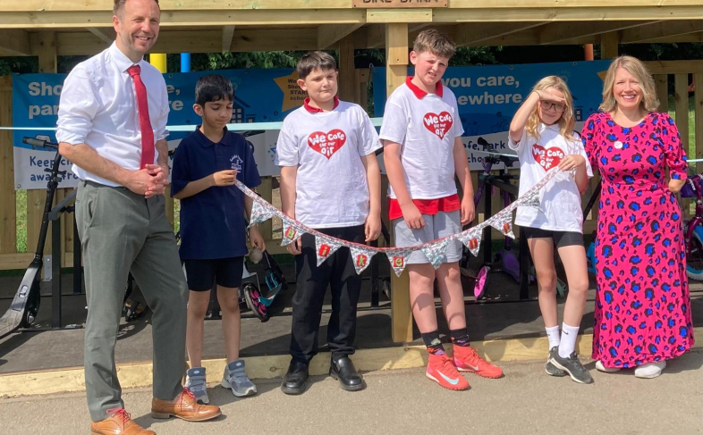A group of adults and school children holding bunting standing in front of a new wooden Bike Barn on their school playground.