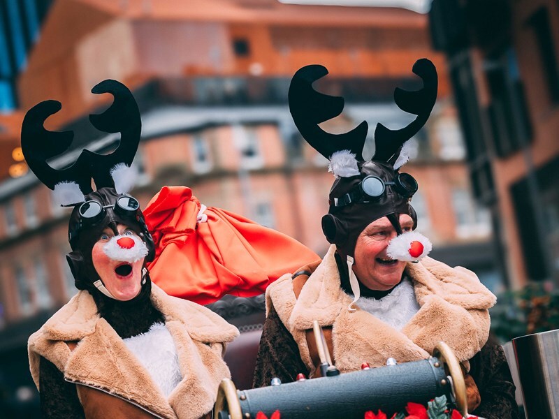 Two people dressed in reindeer costumes with antler headpieces and fur-trimmed coats.