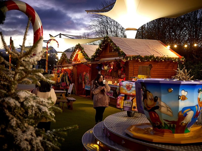 Festive outdoor market scene at dusk with wooden stalls decorated with garlands and lights.