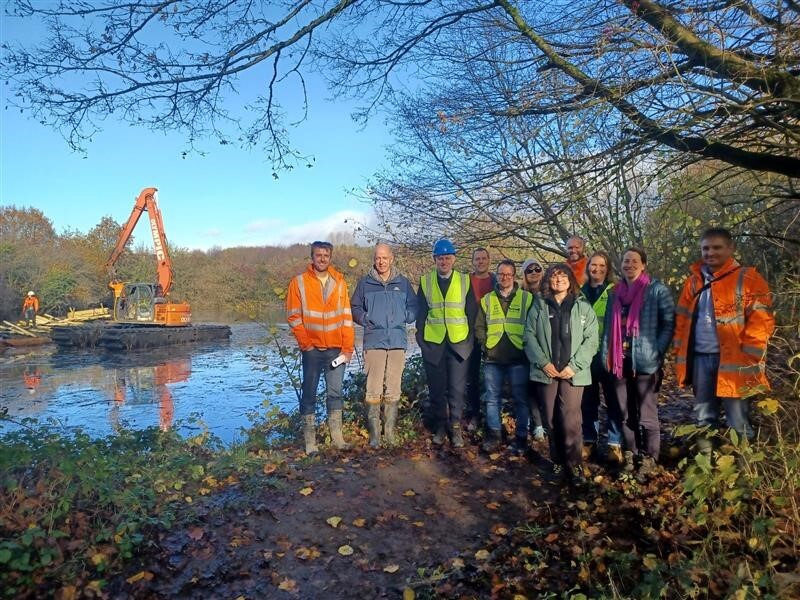 A group of people in high-viz standing at the side of a pond, with a dredger in the background