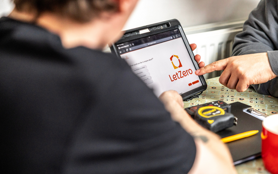 Two people sitting at a table looking at a tablet displaying the Let Zero logo