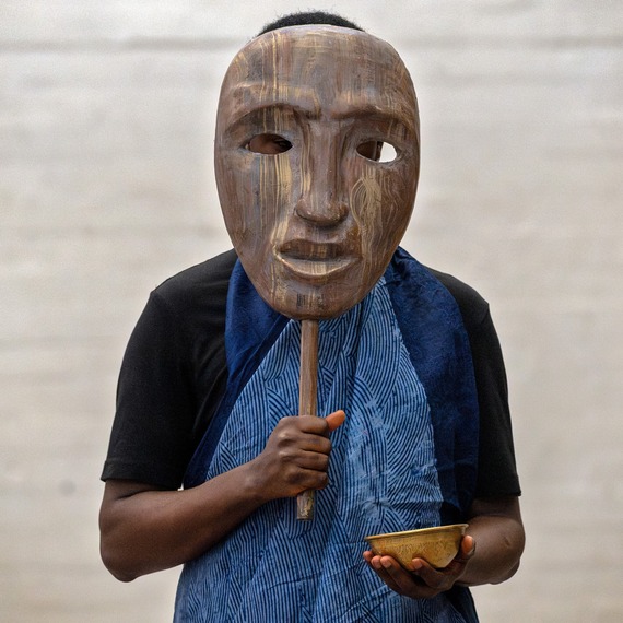 Person stood holding up striking Yoruba mask over their face whilst wearing a vibrant blue gown.