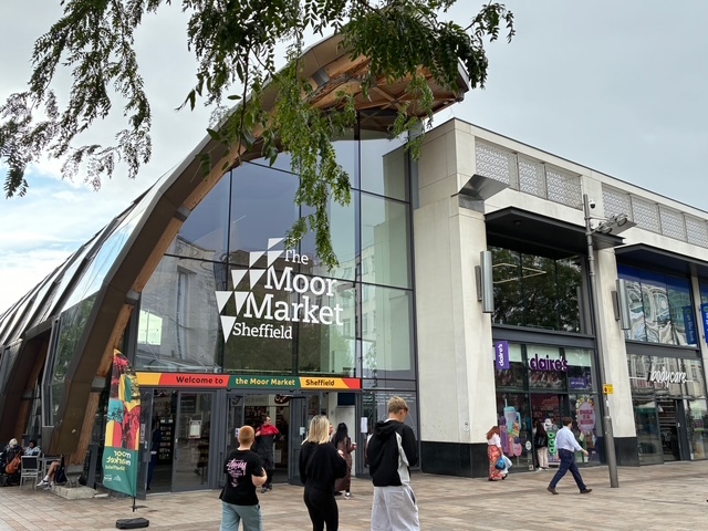 A tree hangs over the top of the photo, with the glass entrance of the Moor Market visible. A number of people walk by.