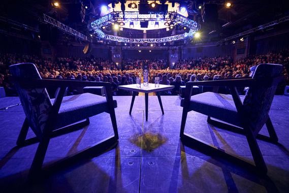 Two empty chairs set up for an interview in front of a packed theatre audience.