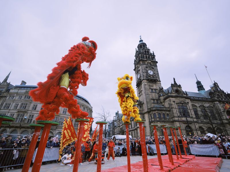 Two performers in vibrant red and yellow dance costumes leap between tall poles during an outdoor celebration.