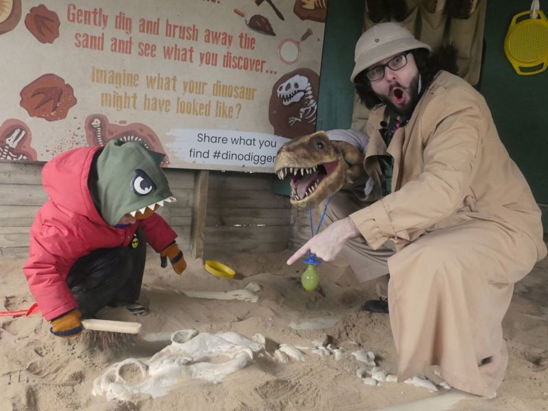 A child in a dinosaur-themed hoodie and a person dressed as an explorer kneel in a sandy dig area, using brushes to uncover replica dinosaur bones.