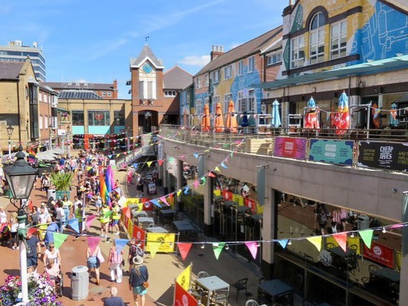 Busy outdoor shopping street decorated with colorful bunting, murals, café seating, and crowds enjoying a sunny day.
