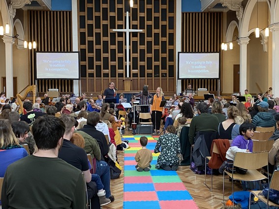 Large audience seated on colourful mats and chairs in a church hall watching musicians and speakers performing at the front beneath a tall cross.