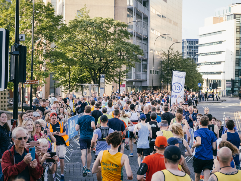 Runners fill a city street during a race, passing spectators and a sign marking the 13‑mile point.