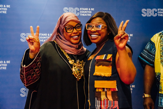 Two people stood in front of a navy media stand posing with two fingers in a peace sign.