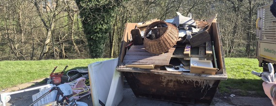 An overflowing yellow skip filled with household item. Further items on the ground around the skip, and grass and trees behind.