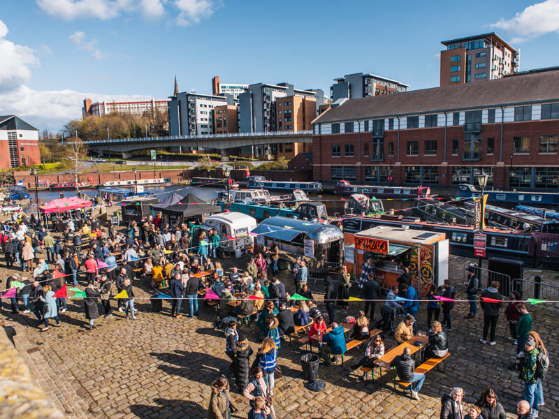 Crowded waterfront market with street food vans, canal boats and picnic tables on a cobbled quay, city buildings in the background.