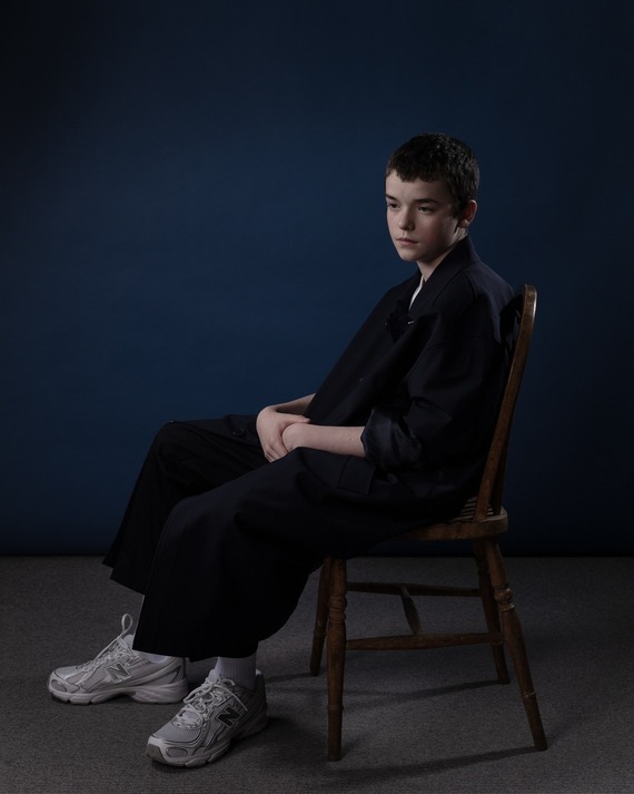 Owen Cooper by Dominic Whisson, 2025. The actor sits in a seat in front of a blue background.