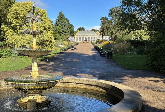 Sheffield Botanical Gardens in the sunshine. There is a blue sky above the Glass Pavilion, with people walking along the path.