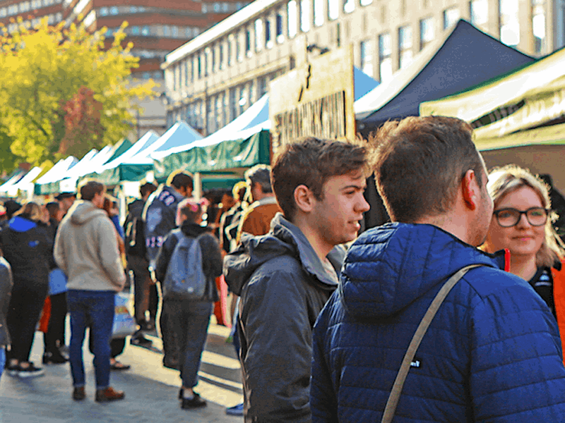 Busy outdoor market with pop-up stalls and people browsing along a city street lined with tents and buildings on a sunny day.