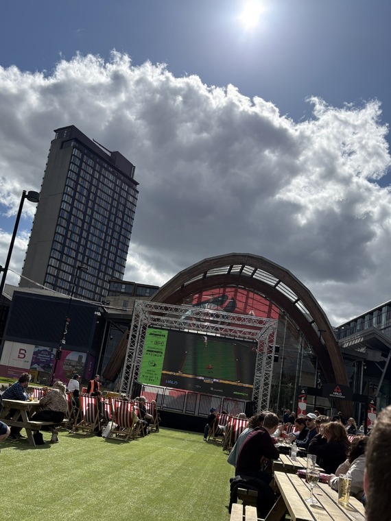 People sit at outdoor tables facing a large screen on Tudor Square, with festival seating, modern buildings, and dramatic clouds overhead.
