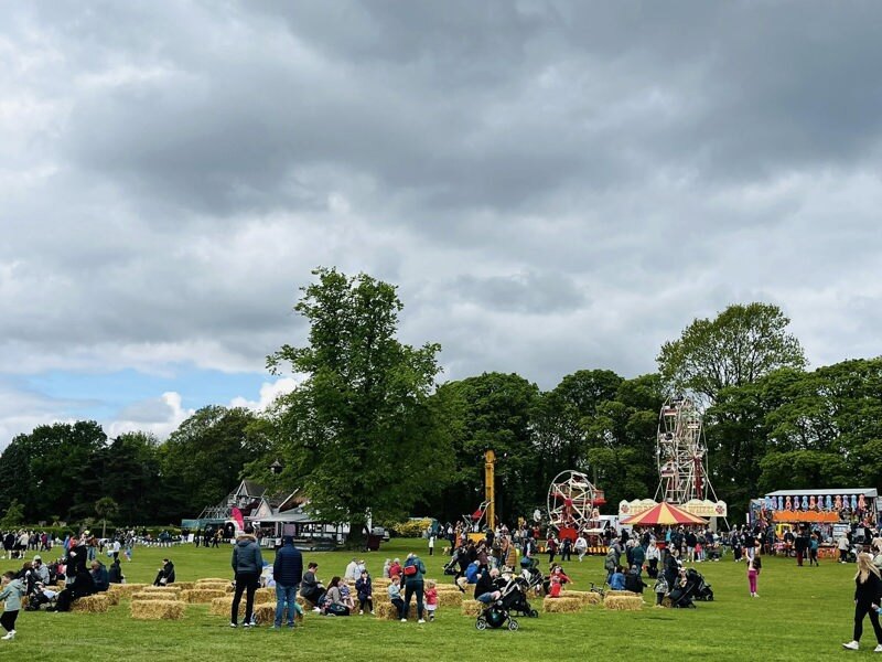 Clouds sit above Graves Park. There is a crowd of people and fairground rides on the Park.