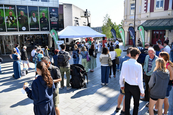 Crowd gathered at an outdoor event with a white marquee and banners in a city square, with people watching a performance.