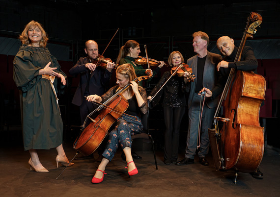 String ensemble on stage with cello, violins, and double bass, alongside a standing performer in a green dress under warm theatre lighting.