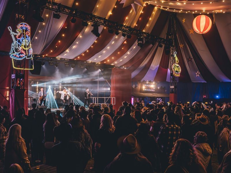 Music is being played on a stage inside a large tent. There is a large crowd and bright lights.