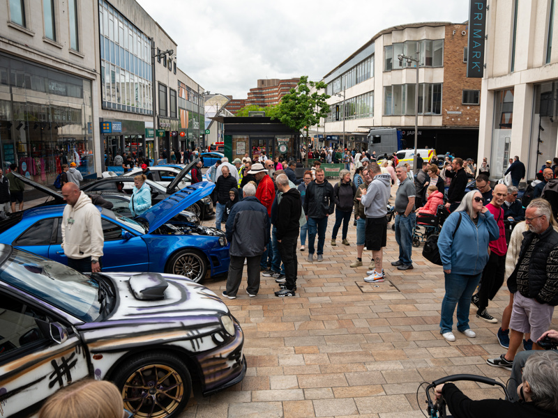People stand looking at a range of classic cars on The Moor. Shops frame the view on both sides of the crowd.