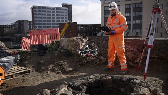 A man in bright orange construction outfit and hard hat flies a drone in a cage above the medieval well at Castlegate