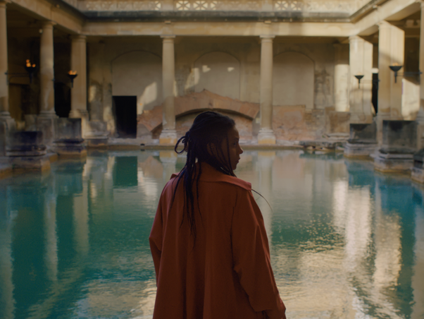 A black woman in her 30s with braided hair is in a semi-silhouette close to us, against a backdrop of water in an ancient temple.