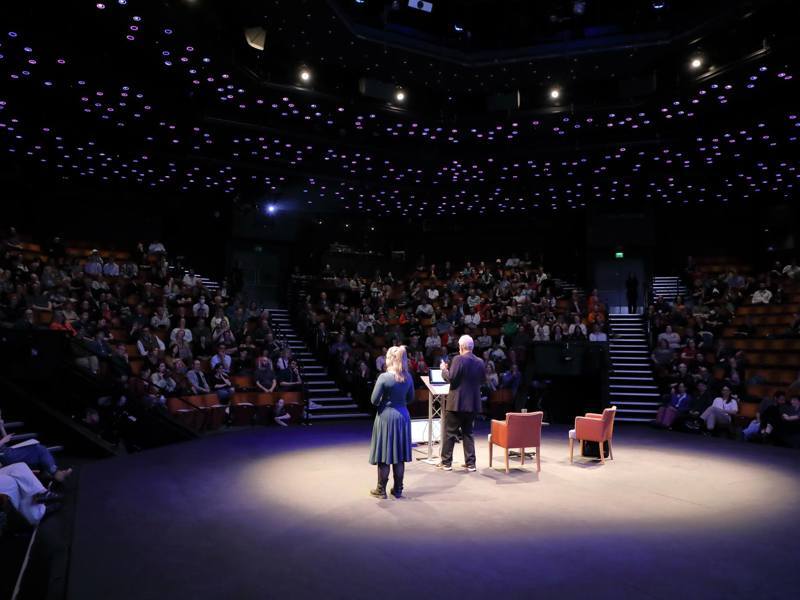 Two speakers present at a podium on a stage in a large auditorium, facing a seated audience under dim blue ceiling lights.