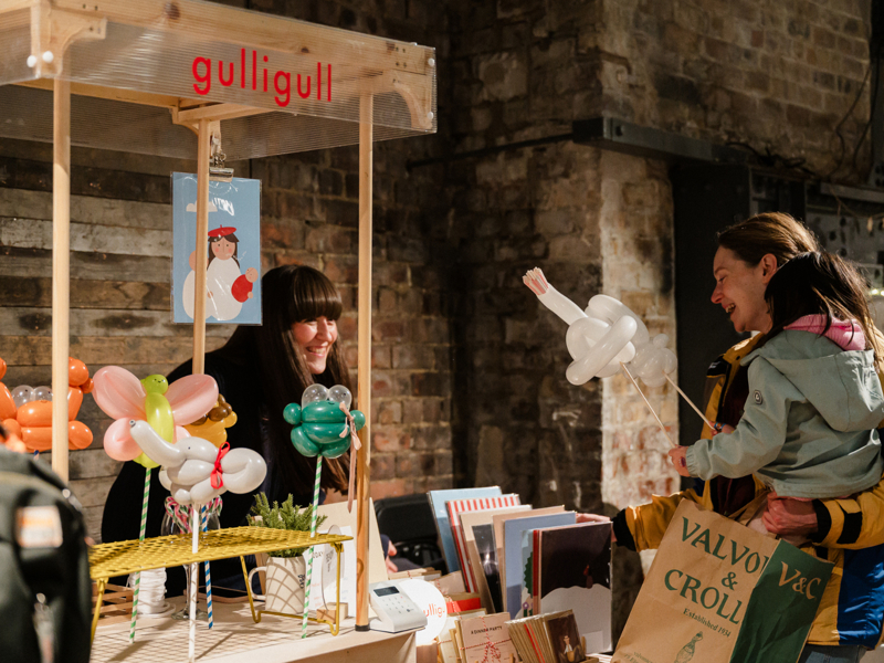 Indoor craft stall “gulligull” with balloon animals and prints; vendor behind table, customer holding balloon sculpture and shopping bag.