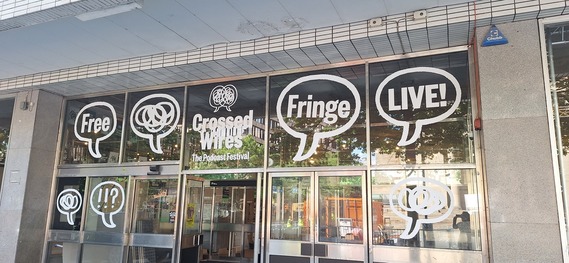 Front of a building with large windows displaying “Crossed Wires: The Podcast Festival” and graphics reading “Free,” “Fringe,” and “Live!”