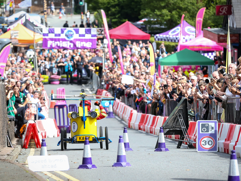 Soapbox race on a city street with crowds behind barriers, a small wheeled vehicle approaching cones, and a “Finish line” banner in the background.
