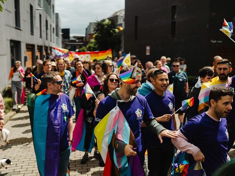 People marching in a Pride parade on a city street, wearing purple shirts, waving rainbow and inclusive flags, with a colorful crowd behind them.