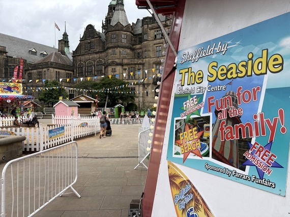 Sign for “Sheffield by the Seaside” beside a temporary beach attraction in Sheffield city centre, with colourful huts, rides and historic buildings.