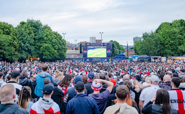 A crowd of England fans watch a big screen on Devonshire Green. Trees frame the screen under a clear sky.