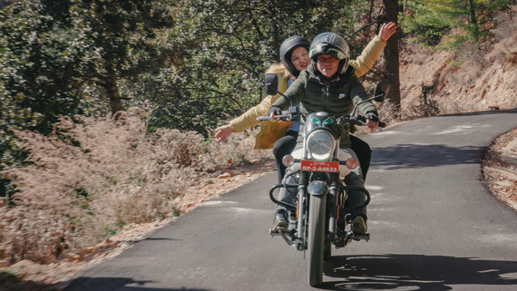 Agent of Happiness - Two people riding a bike in a rural area, the passenger is waving their hands and looking very happy.