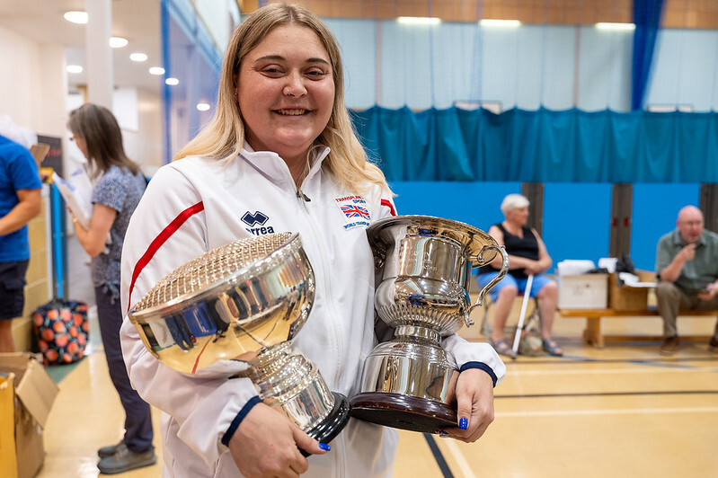Person in a Team GB jacket holding two silver trophies in an indoor sports hall, with spectators and participants in the background.
