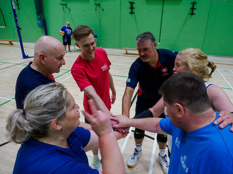 Six people stand in a circle and put their hands together in celebration. They are in a sports hall.