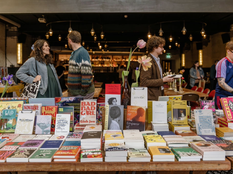 Four people stand around rows of books in a stall. The covers are facing forward.