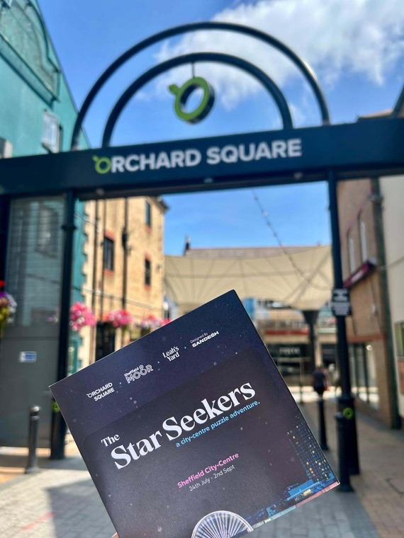 A leaflet with the title 'Star Seekers' on it, in front of Orchard Square entrance, blurred in the background. A blue sky sits above the area.