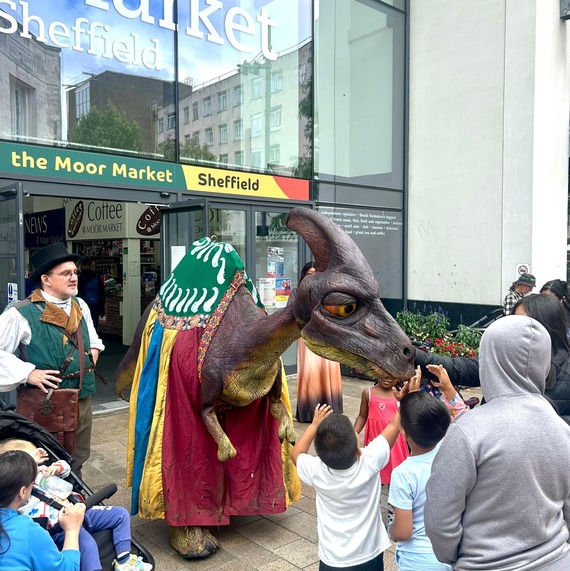 A Dinosaur figure outside the entrance of Moor Market, with kids stroking its head. An assistant worker stands next to the dinosaur.
