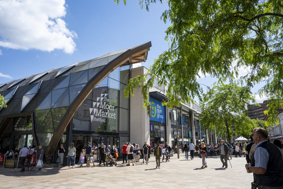 A view of the Moor Market from The Moor. There are people walking past the glass facade, as well as a large overhanging tree.