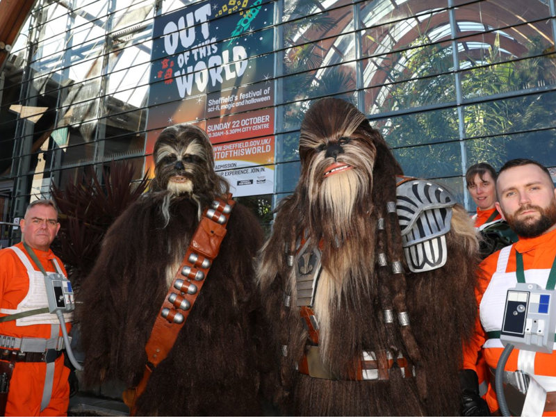 Two people in Star Wars outfits, with three people surrounding them in orange, fighter pilot outfits. They are stood in front of the Winter Garden.