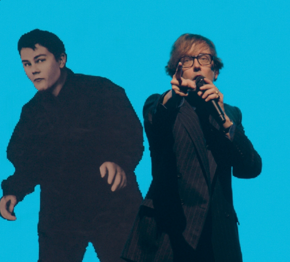 Two men stood in front of a light blue background. On the right is Jarvis Cocker holding a microphone to his face and pointing his finger forward.