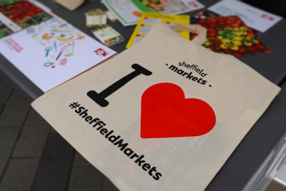 A tote bag that has I heart Sheffield Markets printed on it with a large red love heart. It sits on a table with leaflets surrounding it.