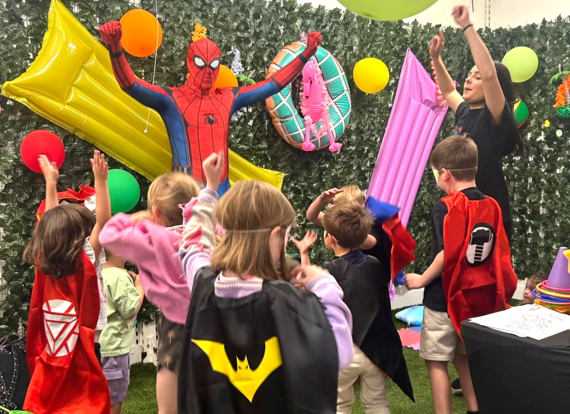 An entertainer dressed up as a superhero at a children's party. The children dance. They all wear superhero capes and masks. 