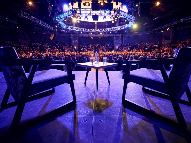 Two chairs in front of a table, facing a huge audience. A blue spotlight shines on the stage with heads looking towards it.